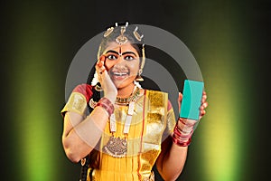Portrait shot of Excited happy Bharatnatyam dancer showing green screen mobile phone by looking at camera on stage -