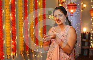 portrait shot of Happy Indian young woman looking at camera by holding diwali diya lamp at home - concept of Traditional