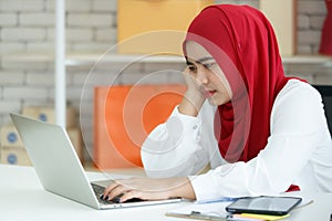 Portrait shot of young Muslim woman wearing a red hijab sitting on a black office chair, looking at the laptop with serious