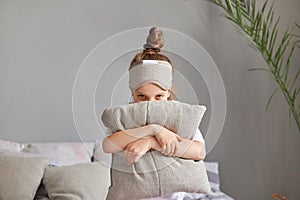 Portrait of shy cute little girl with bun hairstyle sitting in bedroom on bed with pillow hiding her face wearing sleeping mask