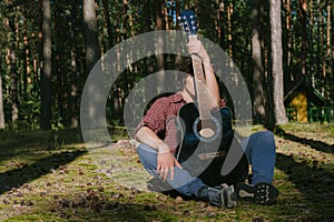 Portrait of a shy guy sitting in the forest, covering his face with a guitar. Against the background of the forest camp