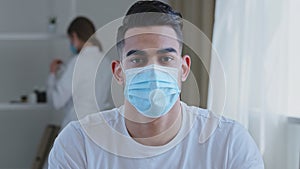 Portrait of sick ethnic arabic man male patient wears medical protective mask on face looking at camera sitting at table