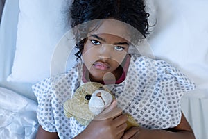 Portrait of sick mixed race girl lying in hospital bed holding teddy bear