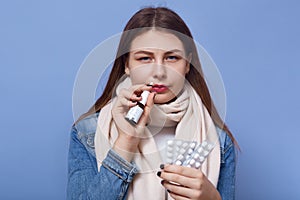 Portrait of sick suffering female standing isolated over blue background in studio, holding nasal drops and pills in both hands,