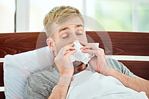 Portrait of a sick young man with runny nose suffering from cold or flu and sneezing while lying in bed at home. Virus
