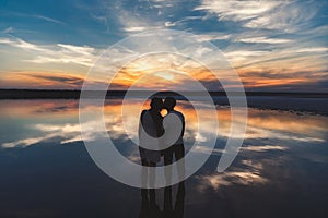 Portrait, silhouette of happy couple watching the colourful bright sunset standing in large lake and kissing, reflection in the wa