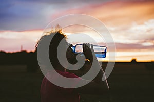 Portrait of a silhouette of a young tourist woman drinking water from a transparent blue bottle in a field, jars at sunset. The gi