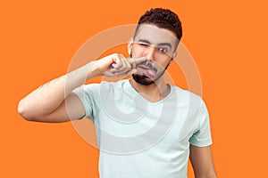 Portrait of silly childish brunette man with beard in casual white t-shirt picking his nose. indoor studio shot isolated on orange