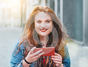 Portrait of sincerely smiling red curled long hair caucasian teen girl walking on the street and video chatting using the modern s