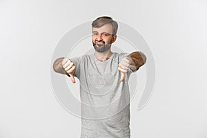 Portrait of skeptical bearded man in casual t-shirt, showing thumbs-down and looking disappointed, disapprove something