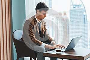 Skilled businessman working with laptop near window with skyscraper. Ornamented.