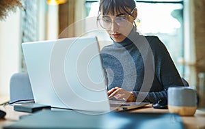 portrait of skilled young female developer in eyeglasses concentrating on screen and typing on laptop while sitting at