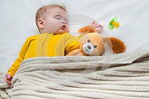 Portrait of a sleeping baby in bed under a blanket with a toy and pacifier nearby