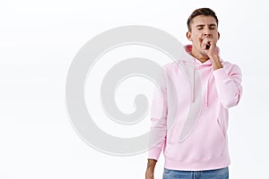 Portrait of sleepy handsome blond man in pink hoodie, close eyes and yawning over fatigue, tired of working late, waking