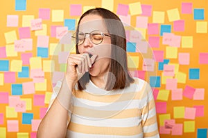 Portrait of sleepy overworked attractive woman wearing striped T-shirt standing against yellow wall covered with stickers, yawning