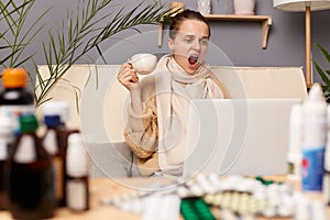 Portrait of sleepy sick unhealthy Caucasian young adult woman wearing warm sweater and scarf sitting on sofa in home interior