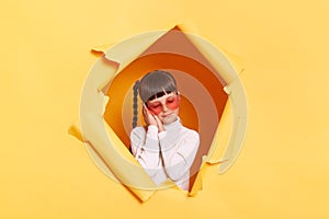 Portrait of sleepy tired little girl with braids and in glasses wearing casual shirt looking through torn hole in yellow paper,
