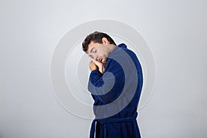 Portrait of sleepy young man wearing blue bathrobe holding hand to forehead, eyes closed feel tired needs to sleep isolated on