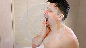 Portrait sleepy young man yawning and looking at mirror in bathroom in morning.