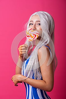 Portrait of a slender woman in a blonde wig posing with a lollipop on a pink background. Cute girl with blue eyes in a