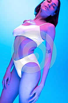 Portrait of slim beautiful young woman posing in underwear isolated over blue background in neon light.