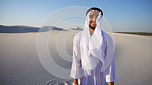 Portrait of slow motion confident male Arabian UAE Sheikh in Kandura in midst of white desert on clear day.
