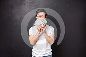 Portrait of sly happy and pleased asian young man winning big cash prize money, hiding face behind fan of dollars