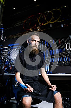 Portrait of small business owner of young man with beard. Guy bicycle mechanic workshop worker sitting with tool in his hand in a