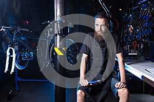 Portrait of small business owner of young man with beard. Guy bicycle mechanic workshop worker sitting with tool in his hand in a