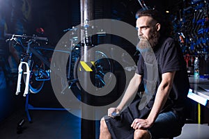 Portrait of small business owner of young man with beard. Guy bicycle mechanic workshop worker sitting with tool in his hand in a