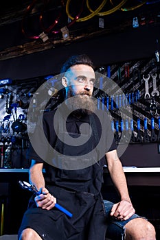 Portrait of small business owner of young man with beard. Guy bicycle mechanic workshop worker sitting with tool in his hand in a