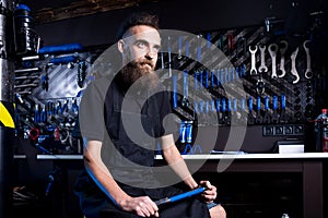 Portrait of small business owner of young man with beard. Guy bicycle mechanic workshop worker sitting with tool in his hand in a