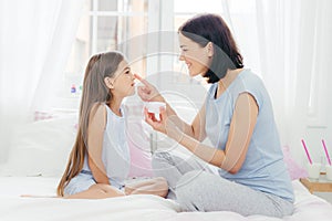 Portrait of small child looks happily at mother who applies cream on her nose, dressed in nightwear, have beauty procedures