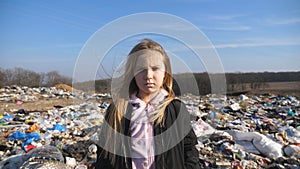 Portrait of small female child looks into camera against the blurred background of dirty garbage dump. Little serious