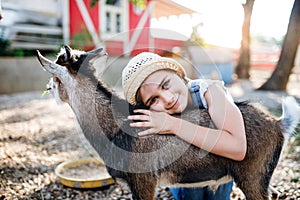 A portrait of small girl outdoors on family animal farm, hugging goat.