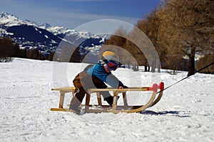 Portrait of small smiling boy sitting on sled on snowy slope on background of sunny mountains Alps and blue sky