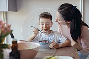 Portrait of small teen boy eating cereals with milk in kichen and mother looking with love.