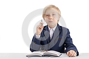 Portrait of smart blond schoolboy in school uniform. Student sits at table with book and pen in his hands. Education concept.