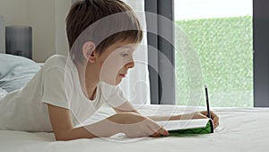 Portrait of smart boy lying in bed at morning and doing homework in notebook. Concept of education, child development.