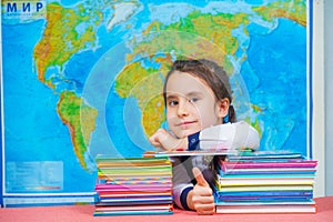 Portrait of a smart cute girl hugging a lot of books on the background of the world map. Thumbs up