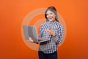 Portrait of smart positive woman with brown hair in checkered casual shirt and denim standing pointing at laptop in her hands,