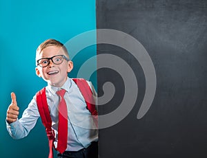 Portrait of smart school child with thumb up near blackboard blank and looking at camera. Happy kid boy in uniform. Go