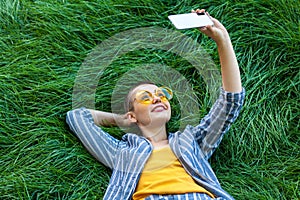 Portrait of smiley young short hair woman in casual blue striped suit, yellow shirt, glasses lying down on green grass holding her