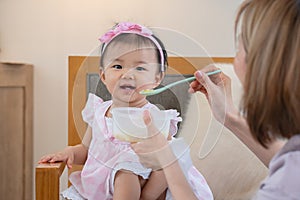 Portrait of smiling adorable Asian baby eating food from spoon, mother feeding little daughter puree or solid foods, toddler