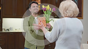 Portrait of smiling adult son talking telling wishes giving bouquet of tulips and gift box to senior mother. Joyful