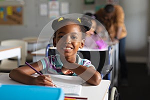 Portrait of smiling african american elementary girl studying while sitting on wheelchair at desk