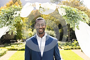 Portrait of smiling african american groom standing under wedding arch in sunny garden