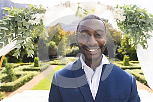 Portrait of smiling african american groom standing under wedding arch in sunny garden