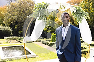 Portrait of smiling african american groom standing under wedding arch in sunny garden, copy space