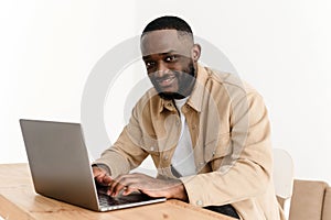 Portrait of smiling african american man looking at camera while sitting at table in home office and working on laptop